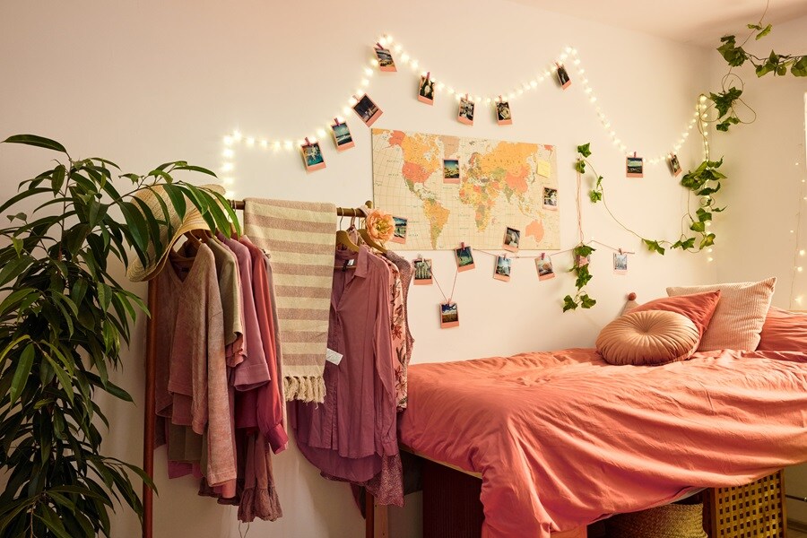 Cozy bedroom with a neatly made bed, pillows, and a green throw. Art prints hang above the bed with Command Strips. A desk, chair, and photos with Command Hooks add charm. Plants enhance warmth.

